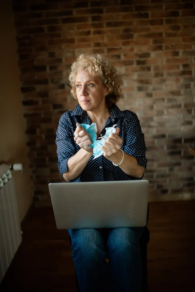 sad facial expression photo — Woman sitting on a chair with a laptop on her lap and tearing apart her face mas